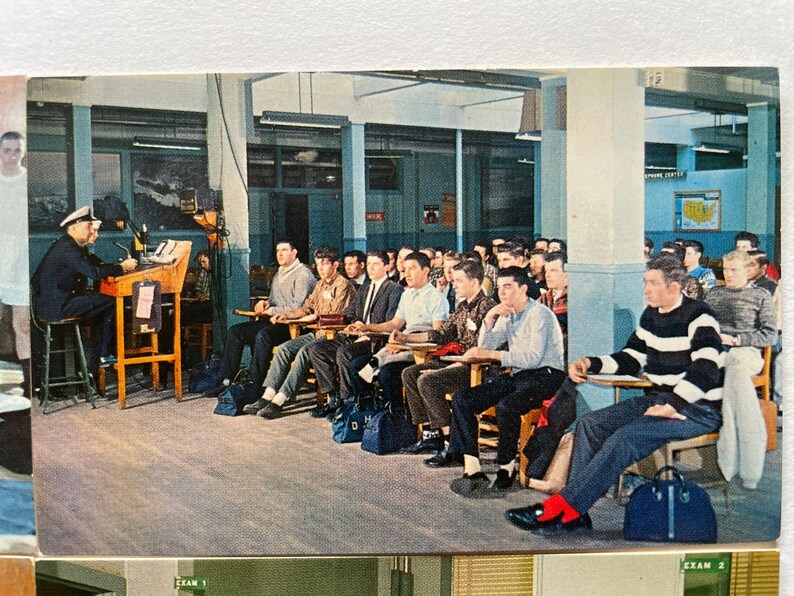 May include: A classroom scene with a teacher in a navy blue uniform standing at a desk with a microphone. Students are seated in rows of desks, listening to the teacher. The classroom has a light blue and white color scheme.