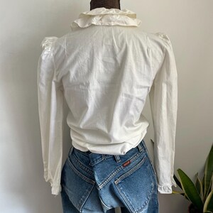 Puede incluir: Blusa blanca de manga larga con cuello de volantes y hombros abullonados. La blusa est&aacute; hecha de una tela ligera y tiene un corte holgado.