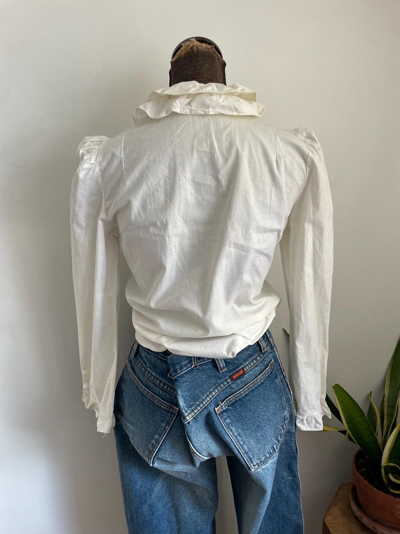 Puede incluir: Blusa blanca de manga larga con cuello de volantes y hombros abullonados. La blusa est&aacute; hecha de una tela ligera y tiene un corte holgado.