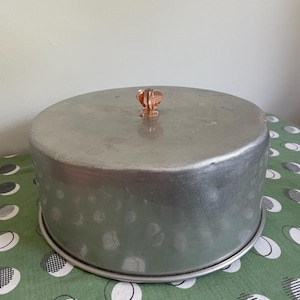 May include: A vintage aluminum cake carrier with a clear, decorative knob. The round carrier has a lid and a base, designed to protect and transport baked goods. The carrier sits on a green tablecloth with a white pattern.