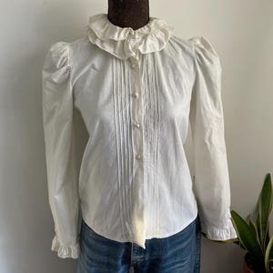 Puede incluir: Una blusa blanca de manga larga con cuello de volantes y cierre de botones. La blusa tiene mangas abullonadas y un corpi&ntilde;o ajustado.