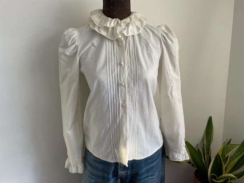 Puede incluir: Una blusa blanca de manga larga con cuello de volantes y cierre de botones. La blusa tiene mangas abullonadas y un corpi&ntilde;o ajustado.