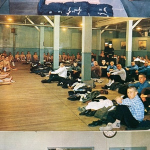 May include: A black and white photograph of a room full of men. The men are sitting on the floor and some are standing. The room has a wooden floor and white walls. There are several windows in the room.