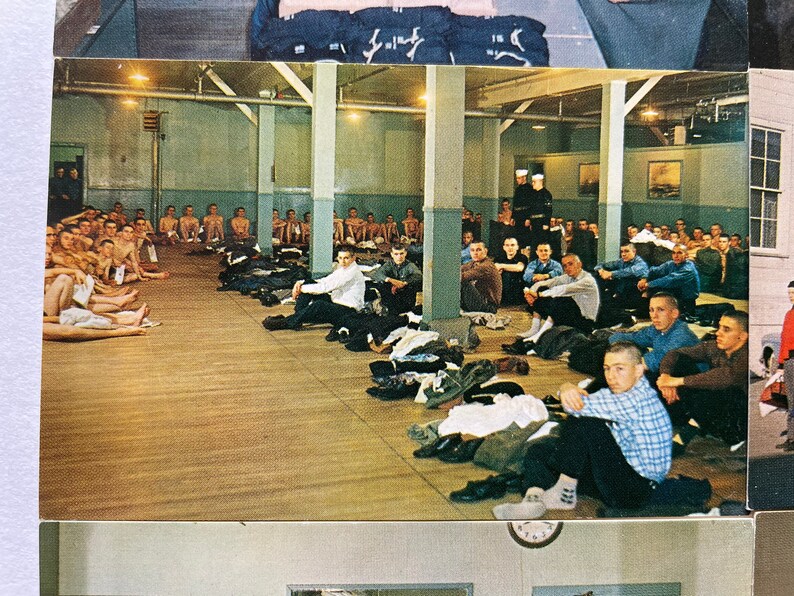 May include: A black and white photograph of a room full of men. The men are sitting on the floor and some are standing. The room has a wooden floor and white walls. There are several windows in the room.