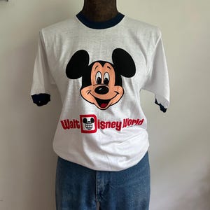 Op de afbeelding: Een wit T-shirt met korte mouwen en donkerblauwe boorden bij de kraag en mouwen. De voorkant toont een afbeelding van het gezicht van Mickey Mouse boven de rode tekst "Walt Disney World" met een klein wereldbolicoon in de letter D.