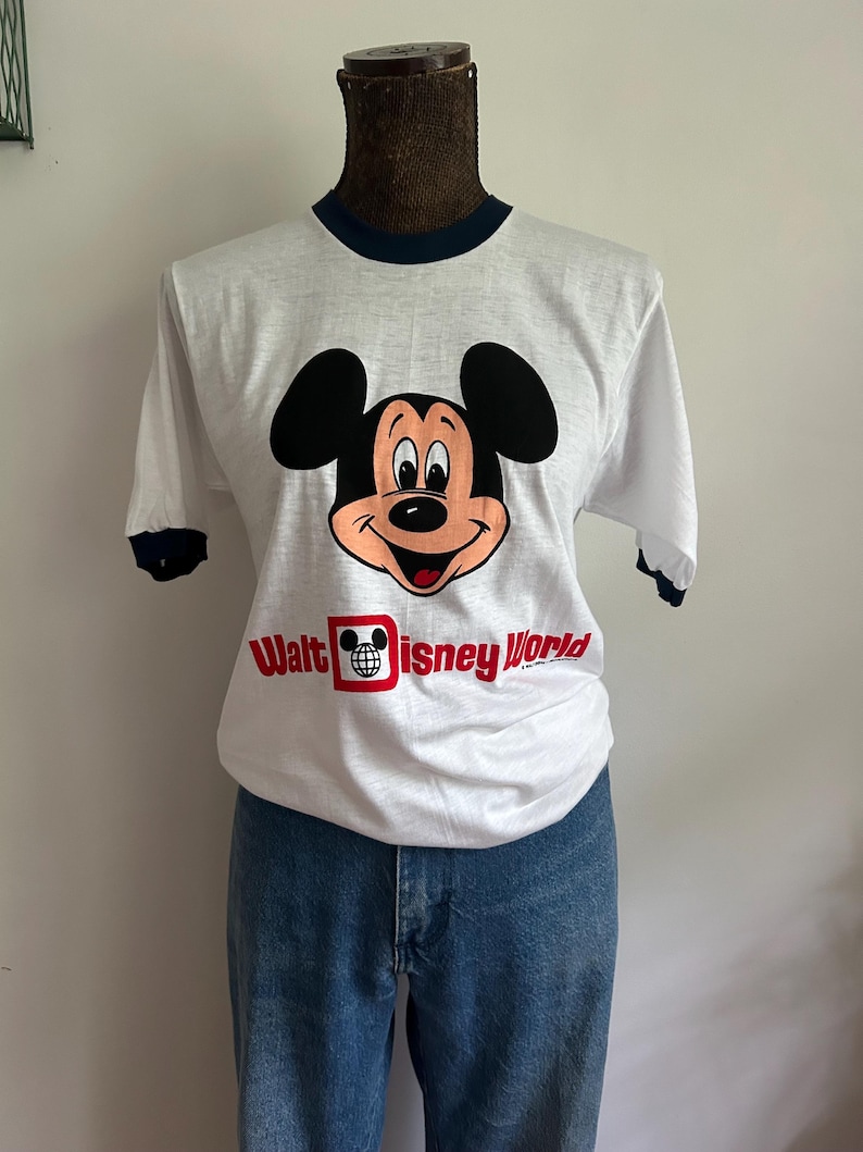 Op de afbeelding: Een wit T-shirt met korte mouwen en donkerblauwe boorden bij de kraag en mouwen. De voorkant toont een afbeelding van het gezicht van Mickey Mouse boven de rode tekst "Walt Disney World" met een klein wereldbolicoon in de letter D.