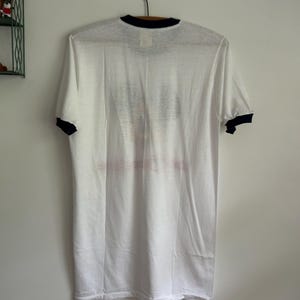 Op de afbeelding: Een wit t-shirt met korte mouwen en een zwarte ronde hals en zwarte mouwboorden. Het kledingstuk heeft een vervaagde grafische print op het voorpand, met een onduidelijke afbeelding en tekst die te wazig is om te kunnen lezen.
