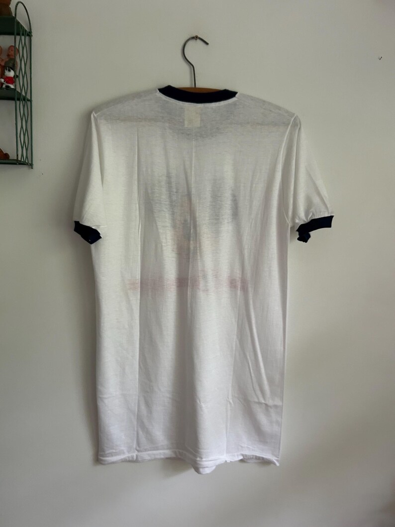 Op de afbeelding: Een wit t-shirt met korte mouwen en een zwarte ronde hals en zwarte mouwboorden. Het kledingstuk heeft een vervaagde grafische print op het voorpand, met een onduidelijke afbeelding en tekst die te wazig is om te kunnen lezen.