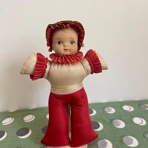 May include: A vintage doll with a painted face, wearing a cream-colored top with red ruffled trim and matching red pants. The doll has a brown bonnet and is standing on a round, light-colored base. The doll's arms are outstretched.