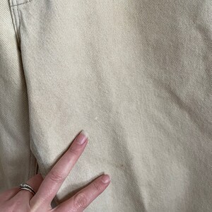 May include: Close-up of a pair of light beige denim pants. The fabric has a subtle texture and a slight sheen.