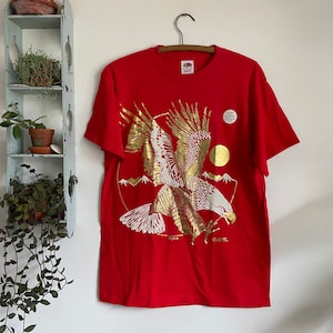May include: Red t-shirt with a gold foil print of an eagle in flight. The eagle is flying over a mountain range and a sun. The shirt has a tag that says "ROSE."
