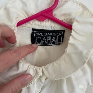Puede incluir: Blusa blanca de cuello con cuello de volantes y un bot&oacute;n de perla. La blusa tiene una etiqueta negra que dice "Diane Gilman for Cabal".