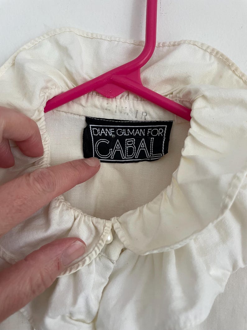 Puede incluir: Blusa blanca de cuello con cuello de volantes y un bot&oacute;n de perla. La blusa tiene una etiqueta negra que dice "Diane Gilman for Cabal".