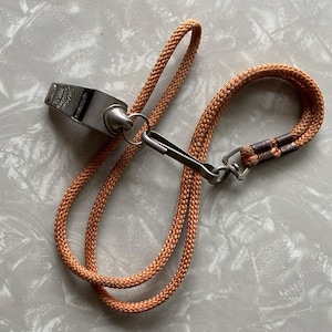 May include: A silver metal whistle with an orange braided lanyard. The whistle has text on its surface and is connected to the lanyard with a metal clasp. The lanyard is made of a thick, durable material.