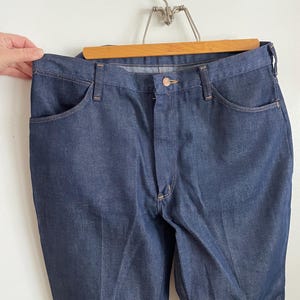 May include: Dark blue denim jeans hanging on a wooden hanger. The jeans have a classic five-pocket design with a button and zipper closure. The denim appears to be a medium weight, with a slight sheen. The jeans are displayed against a white background.