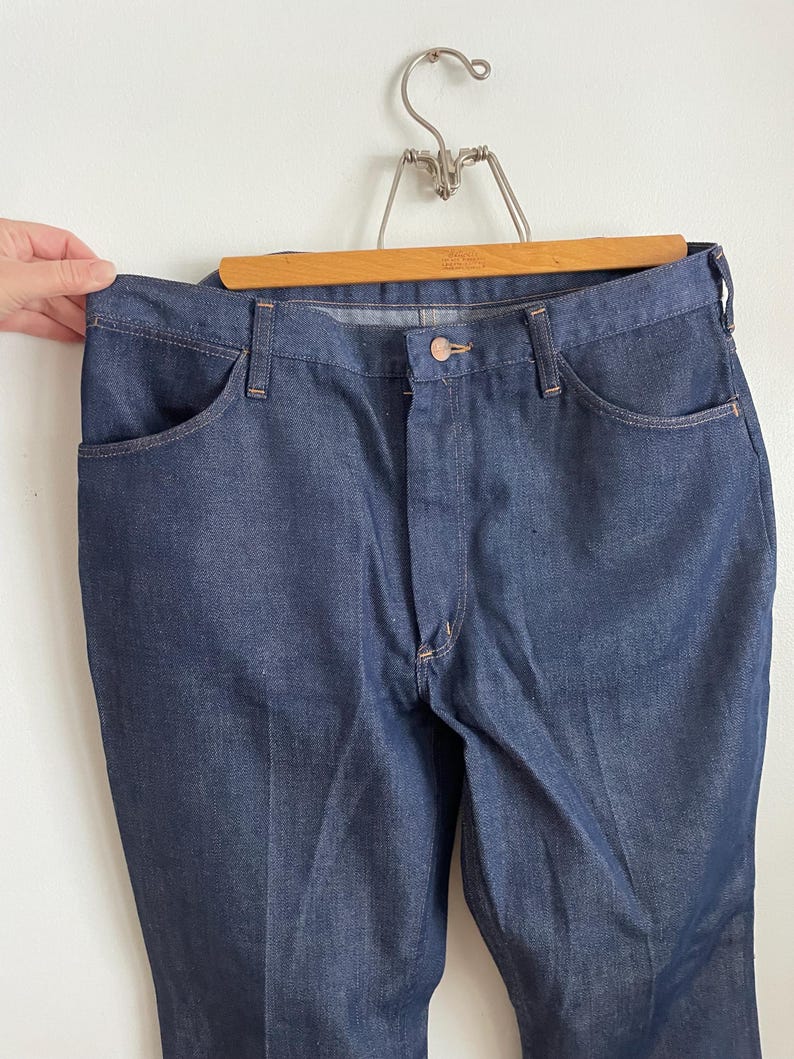 May include: Dark blue denim jeans hanging on a wooden hanger. The jeans have a classic five-pocket design with a button and zipper closure. The denim appears to be a medium weight, with a slight sheen. The jeans are displayed against a white background.