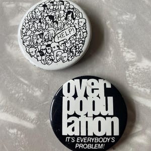 May include: Two round button pins with black and white designs. The first pin has a white background with a black outline of many faces and the word "HELP!" in a speech bubble. The second pin has a black background with white text that reads "over population" and "IT'S EVERYBODY'S PROBLEM!"