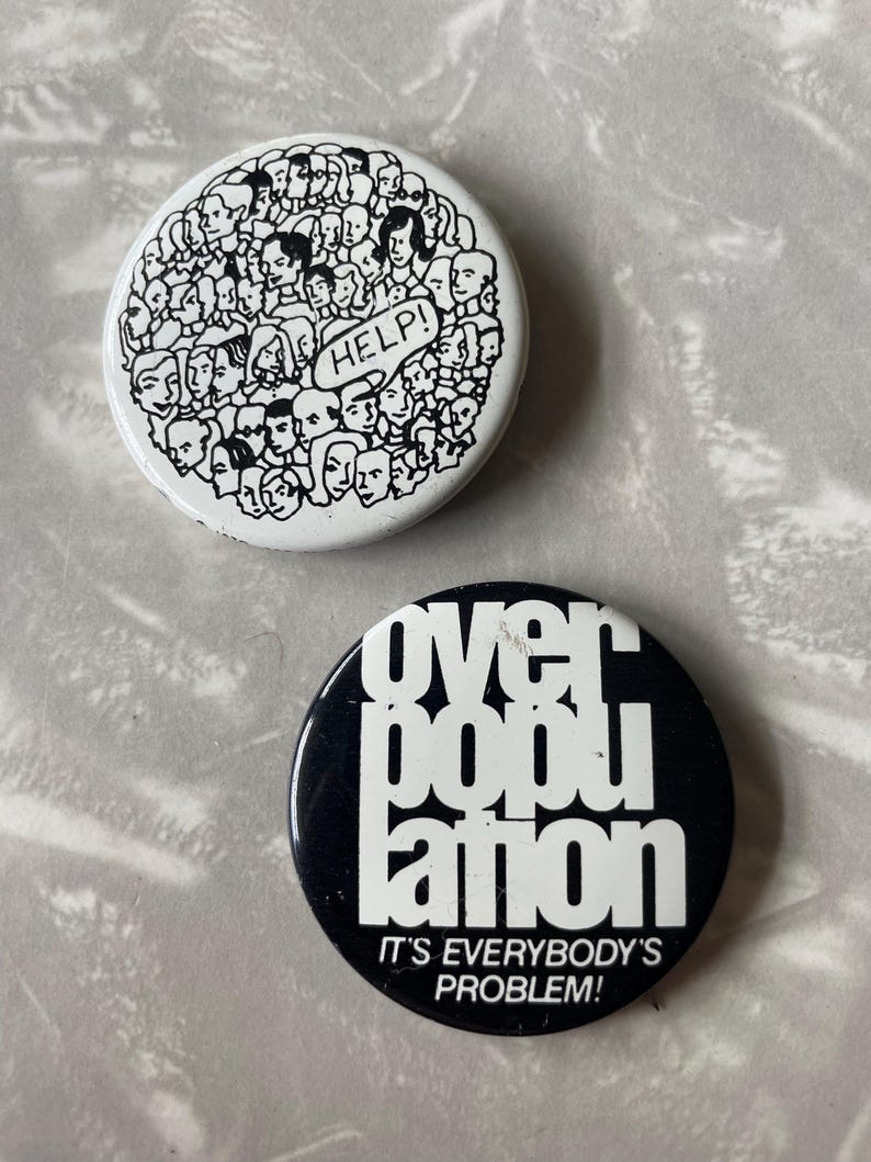 May include: Two round button pins with black and white designs. The first pin has a white background with a black outline of many faces and the word "HELP!" in a speech bubble. The second pin has a black background with white text that reads "over population" and "IT'S EVERYBODY'S PROBLEM!"