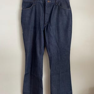 May include: Dark blue denim jeans hanging on a wooden hanger. The jeans have a classic five-pocket design, a button and zipper closure, and a flared leg. The fabric appears to be a medium-weight denim.
