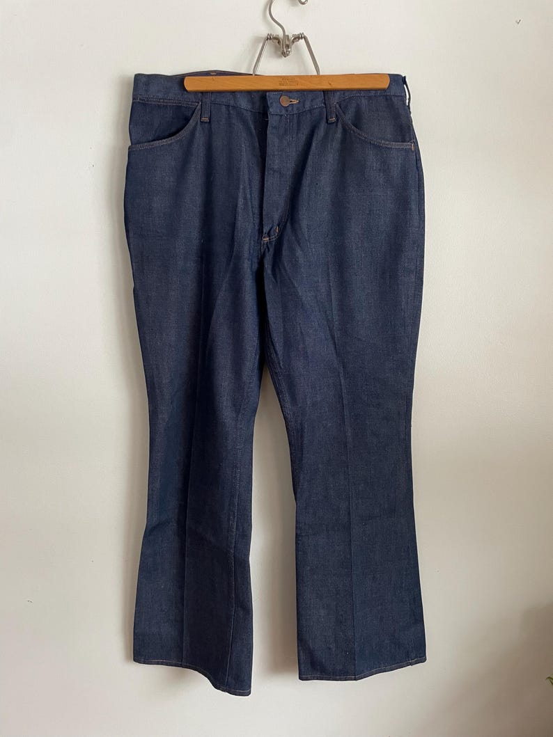 May include: Dark blue denim jeans hanging on a wooden hanger. The jeans have a classic five-pocket design, a button and zipper closure, and a flared leg. The fabric appears to be a medium-weight denim.