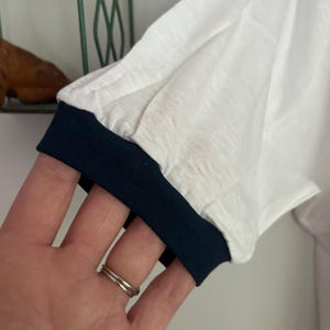 Op de afbeelding: Een wit t-shirt met korte mouwen en een donkerblauwe geribbelde boord aan de mouw. De stof heeft een lichtgewicht, enigszins getextureerd uiterlijk en de mouw is afgewerkt met een strakke zoom in een contrasterende kleur.