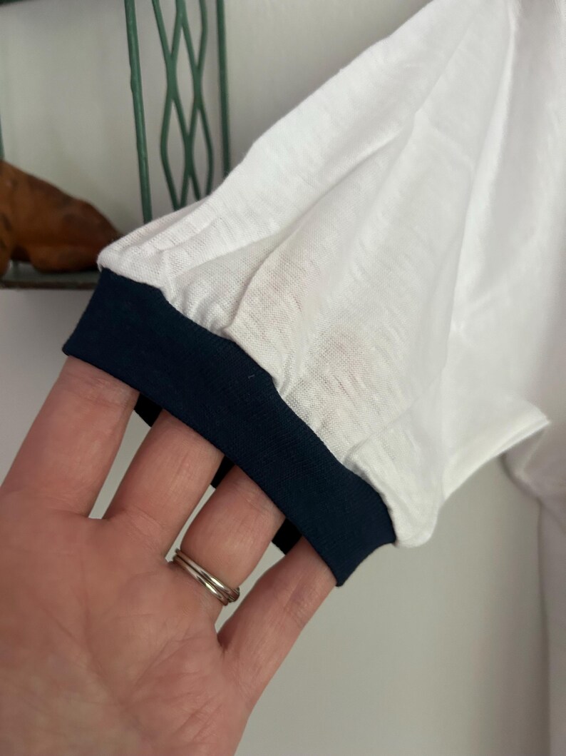 Op de afbeelding: Een wit t-shirt met korte mouwen en een donkerblauwe geribbelde boord aan de mouw. De stof heeft een lichtgewicht, enigszins getextureerd uiterlijk en de mouw is afgewerkt met een strakke zoom in een contrasterende kleur.