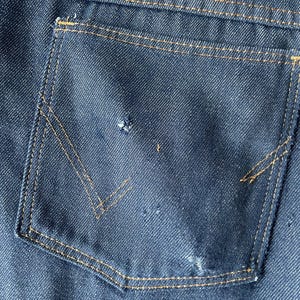 May include: Close-up of a dark blue denim jeans pocket with a V-shaped design. The pocket is outlined with tan stitching. The denim has a textured appearance with some wear and tear.