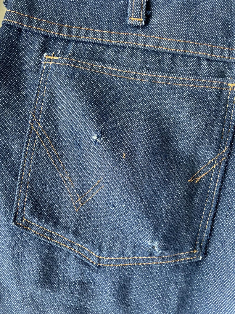 May include: Close-up of a dark blue denim jeans pocket with a V-shaped design. The pocket is outlined with tan stitching. The denim has a textured appearance with some wear and tear.