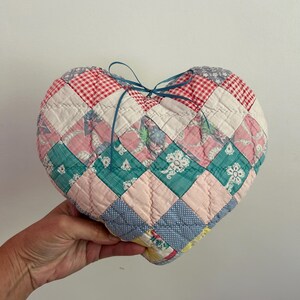 May include: A heart-shaped quilted pillow constructed from various fabric squares in red and white gingham, pink floral patterns, teal, and light blue prints. The pillow features a thin blue ribbon tied in a bow at the top center.