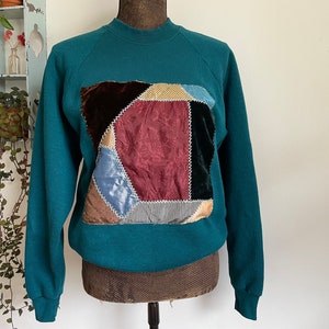 May include: Teal blue sweatshirt with a large patchwork design on the front. The patchwork is made of different colors and textures of fabric, including velvet, corduroy, and striped fabric.