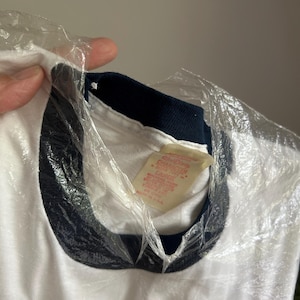 Op de afbeelding: Een wit t-shirt met een donker marineblauwe geribbelde ronde hals. De binnenkant van de kraag heeft een cr&egrave;mekleurig label met rode tekst die Disney Character Fashions, maat Large en Made in U.S.A. vermeldt. Het kledingstuk zit gedeeltelijk in plastic.