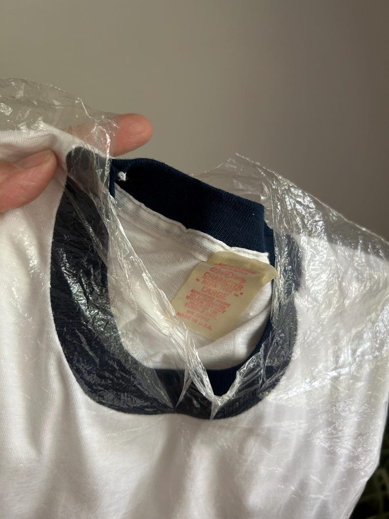 Op de afbeelding: Een wit t-shirt met een donker marineblauwe geribbelde ronde hals. De binnenkant van de kraag heeft een cr&egrave;mekleurig label met rode tekst die Disney Character Fashions, maat Large en Made in U.S.A. vermeldt. Het kledingstuk zit gedeeltelijk in plastic.
