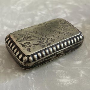 May include: A rectangular silver-toned metal box featuring an engraved design on the lid with geometric triangular patterns and floral motifs. The edges of the lid are decorated with a repeating embossed bead-like border pattern.