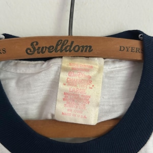 Op de afbeelding: Een wit t-shirt met een donkerblauwe geribbelde kraag hangend aan een houten Swelldom-kledinghanger. Het binnenlabel vermeldt 100% COTTON WASHABLE, WALT DISNEY CHARACTER, LARGE, MACHINE WASH IN LUKEWARM WATER, DO NOT BLEACH OR TUMBLE DRY, RN 12982, MADE IN U.S.A.