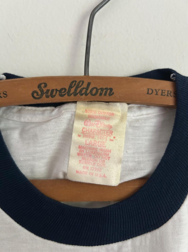 Op de afbeelding: Een wit t-shirt met een donkerblauwe geribbelde kraag hangend aan een houten Swelldom-kledinghanger. Het binnenlabel vermeldt 100% COTTON WASHABLE, WALT DISNEY CHARACTER, LARGE, MACHINE WASH IN LUKEWARM WATER, DO NOT BLEACH OR TUMBLE DRY, RN 12982, MADE IN U.S.A.