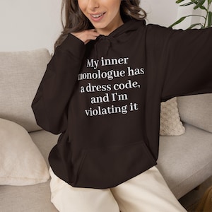 Puede incluir: Una sudadera con capucha de color marrón oscuro con texto blanco impreso en el pecho que dice, My inner monologue has a dress code, and I'm violating it. La prenda incluye un bolsillo tipo canguro frontal, mangas largas y capucha.