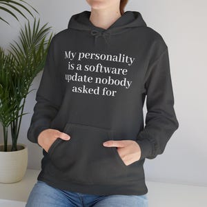 Op de afbeelding: Een donker antracietgrijze hoodie met de witte tekst: My personality is a software update nobody asked for. Het kledingstuk is voorzien van een kangoeroezak aan de voorzijde, lange mouwen met geribbelde boorden en een capuchon met trekkoord.