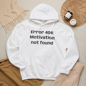 Puede incluir: Una sudadera con capucha blanca que presenta el texto negro "Error 404: Motivation not found" impreso en el frente. La prenda incluye una capucha con cordones blancos, un bolsillo delantero tipo canguro, mangas largas y puños acanalados.