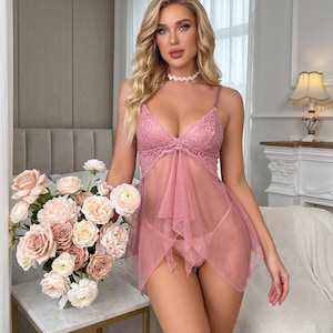 Könnte beinhalten: Ein altrosa Babydoll-Dessous-Set mit einem transparenten Netzrock mit Zipfelsaum und einem spitzenbesetzten Oberteil mit dünnen Spaghettiträgern. Das Set enthält einen passenden Tanga und wird mit einer Perlenhalskette getragen.