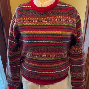 May include: A long-sleeve crew neck sweater featuring a multi-colored horizontal fair isle pattern in shades of red, green, yellow, and dark blue. The sweater has a solid red ribbed collar, cuffs, and hem, worn over a white collared shirt.