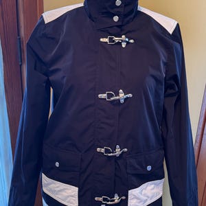 May include: A black jacket featuring white panels on the shoulders and lower front pockets. The garment includes a high collar with two silver-tone snap buttons and four silver-tone metal toggle closures down the center front placket.