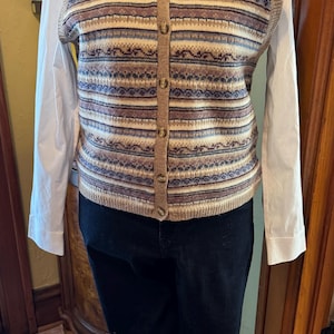 May include: A sleeveless knit button-down vest featuring horizontal stripes in shades of tan, blue, and brown, worn over a white long-sleeved collared shirt and dark pants, paired with brown leather boat shoes with laces and white stitching.
