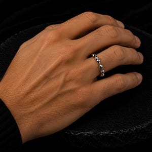 Könnte beinhalten: Ein silberner Ring mit einer Reihe kleiner, runder Perlen wird an einer Hand getragen. Der Ring befindet sich am Ringfinger. Der Hintergrund ist schwarz.