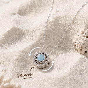 Peut inclure: Un collier pendentif argenté avec un design rotatif circulaire et une pierre bleu clair au centre. La monture en métal présente des détails découpés complexes et une bordure perlée. Le mot spinner est écrit sous le pendentif avec des flèches.