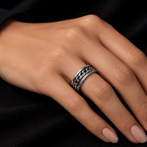 May include: A silver ring with a chain design is displayed on a finger. The ring features a textured band and a rotating chain element. The background is a dark, solid colour.