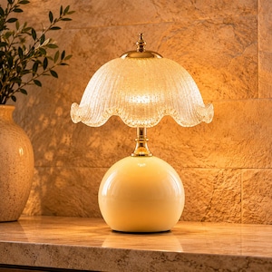 May include: A table lamp featuring a smooth, cream-colored spherical base and a gold-toned metal neck. The lamp is topped with a translucent, textured glass shade that has a scalloped edge and a small gold finial at the very top center.