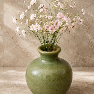 Könnte beinhalten: Eine runde, olivgrüne Keramikvase mit glatter, glasierter Oberfläche und engem Hals, gefüllt mit einem Strauß zarter, hellrosa Blüten an dünnen, verzweigten Stielen. Die Vase hat einen bauchigen Körper und eine schlichte, leicht ausgestellte Öffnung.