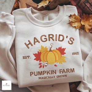 Puede incluir: Una sudadera de cuello redondo color crema con texto bordado en marrón que dice HAGRID'S PUMPKIN FARM EST. 1962 MAGICALLY GROWN. El diseño central incluye una calabaza amarilla bordada rodeada de hojas de otoño rojas y amarillas.