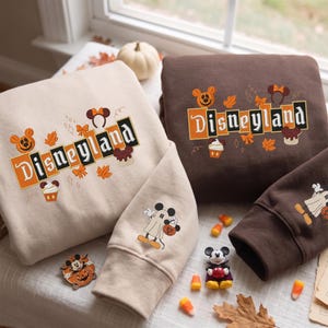 Peut inclure: Un sweat-shirt beige et un marron, chacun portant le texte Disneyland dans une police stylisée. Les motifs incluent des feuilles d'automne orange et marron, des serre-têtes oreilles de Mickey et des cupcakes. Une manche montre un Mickey fantôme.