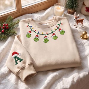 Könnte beinhalten: Ein beigefarbenes Sweatshirt mit Rundhalsausschnitt und einer Stickerei aus grünen Grinch-Gesichtern mit Weihnachtsmützen, die durch eine Lichterkette verbunden sind. Auf dem Ärmel befindet sich ein großer grüner Buchstabe A mit Weihnachtsmütze.
